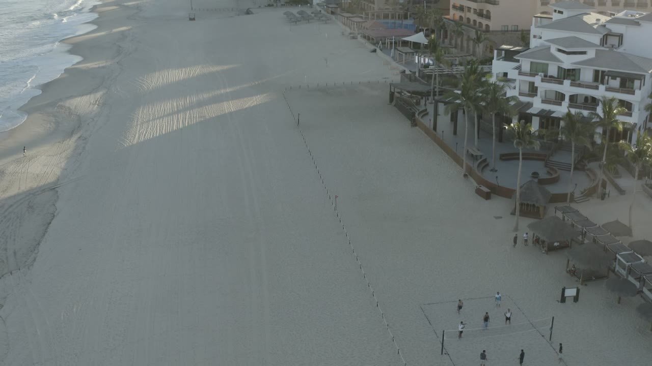 Aerial Los Cabos beach view with rippled sand, a small court near the lower frame, low waves along the shore, palm trees, and white beachfront buildings with no recognizable people.