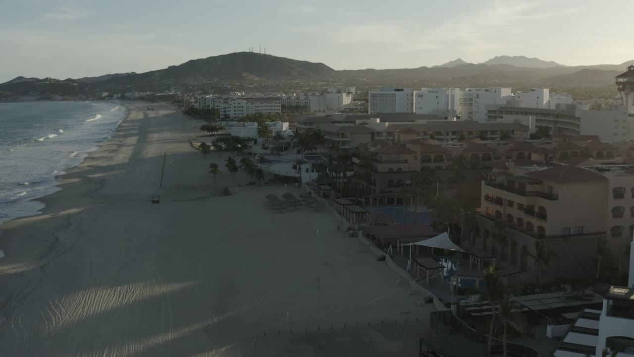 Aerial view of beach, ocean waves, resort buildings, and hills at low sun.