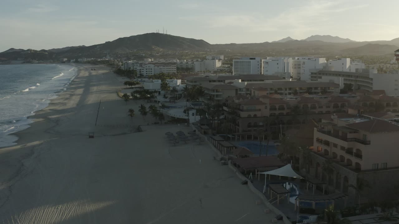 Wide aerial view in Los Cabos showing the shoreline running beside hotel buildings, palm trees, pool areas, mountain silhouettes, and small distant figures on the beach.