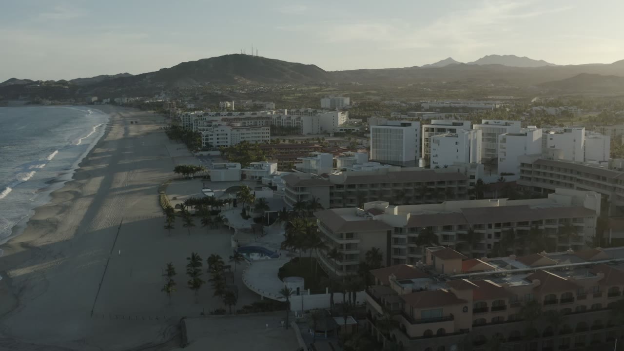 Wide aerial over Los Cabos, Mexico, showing wave lines farther down the beach, a row of white hotel structures, darker foreground resort roofs, distant hills, and no visible people.