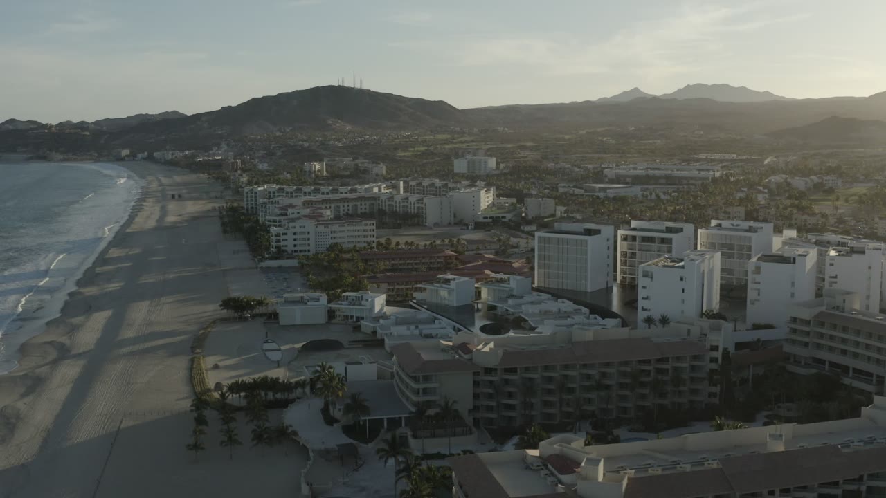 Early frame from Los Cabos, Mexico showing surf and beach on the left, white hotel buildings in the foreground, low city blocks inland, hazy hills beyond, and no visible people.