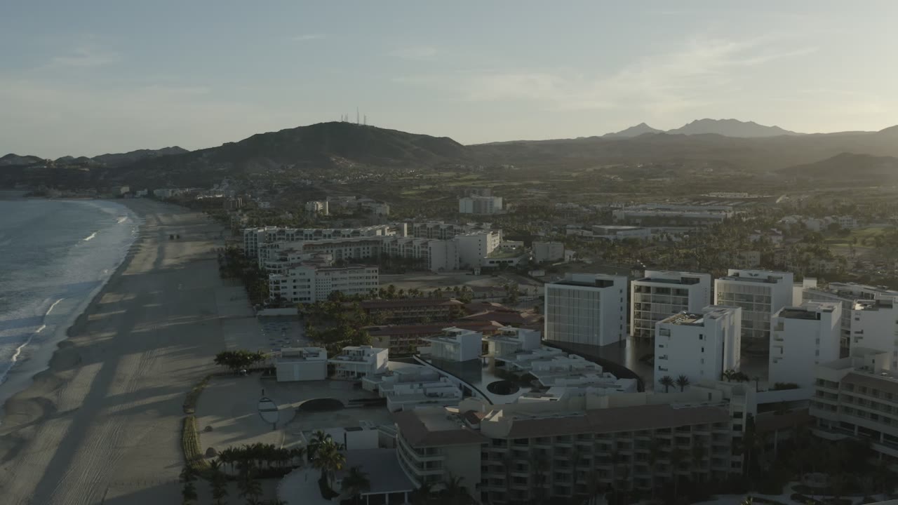 Los Cabos aerial frame with the shoreline curving along the left edge, hotel structures and rooftops below, inland streets and buildings in muted light, mountain ridges in the distance, and no visible people.