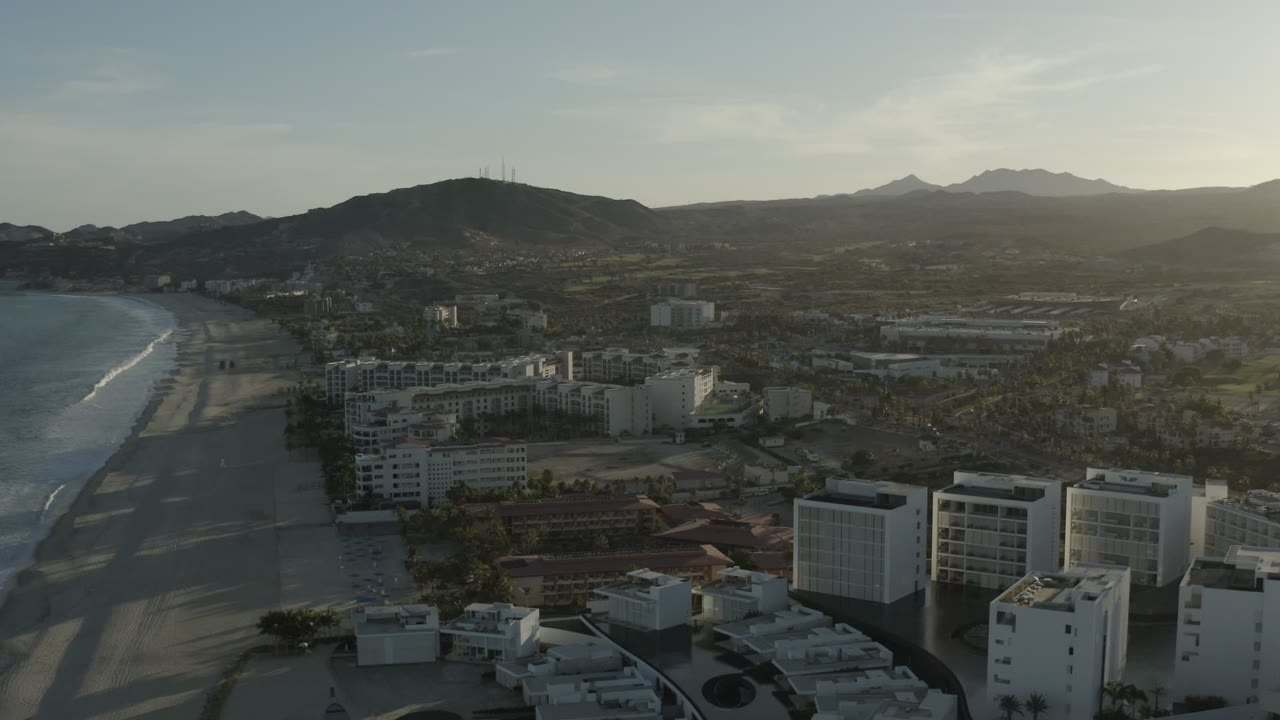 Aerial view of beach, coastal buildings, and mountains at sunset.