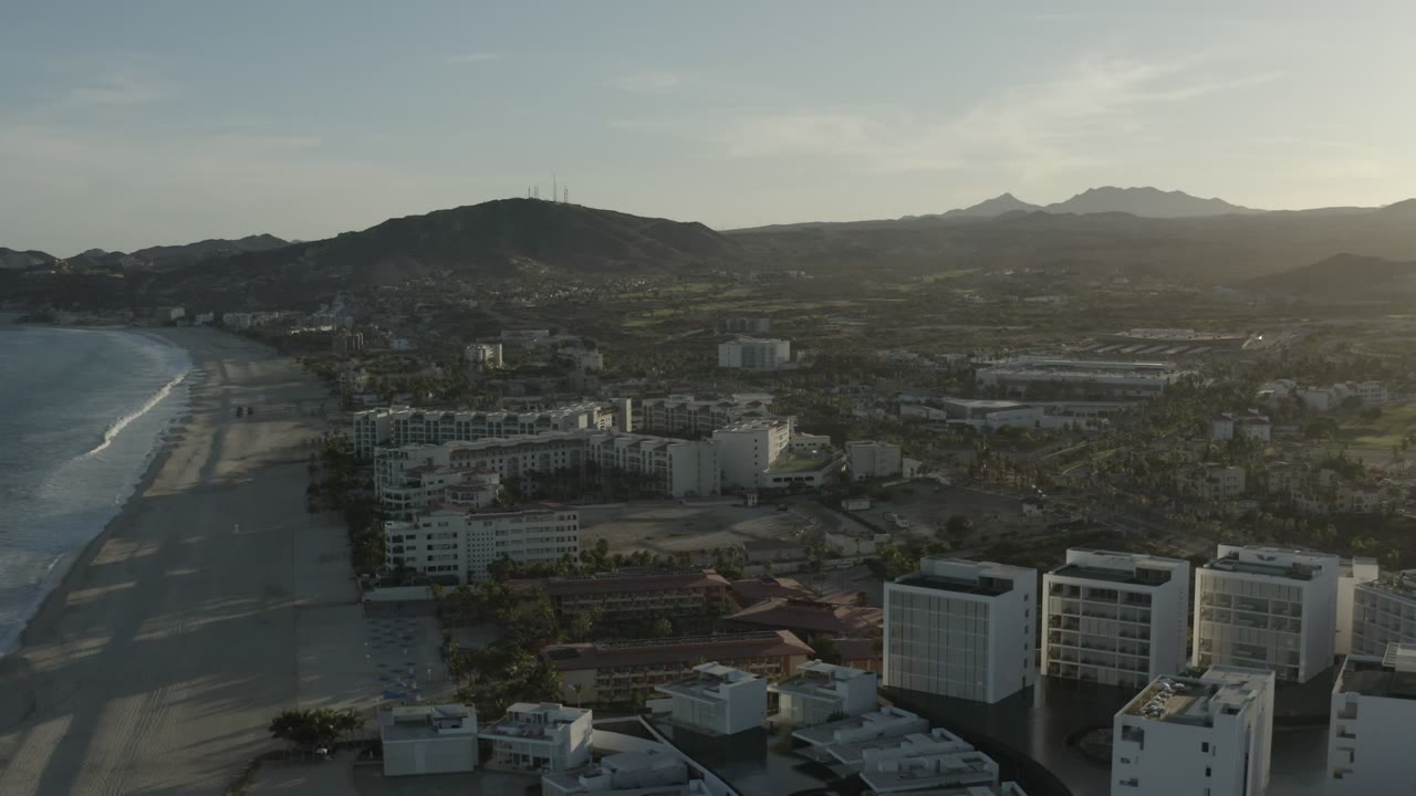 Aerial view over Los Cabos, Mexico, showing the beach curving along the left side, clustered waterfront buildings below, darker hills near center, distant mountains, and no visible people.