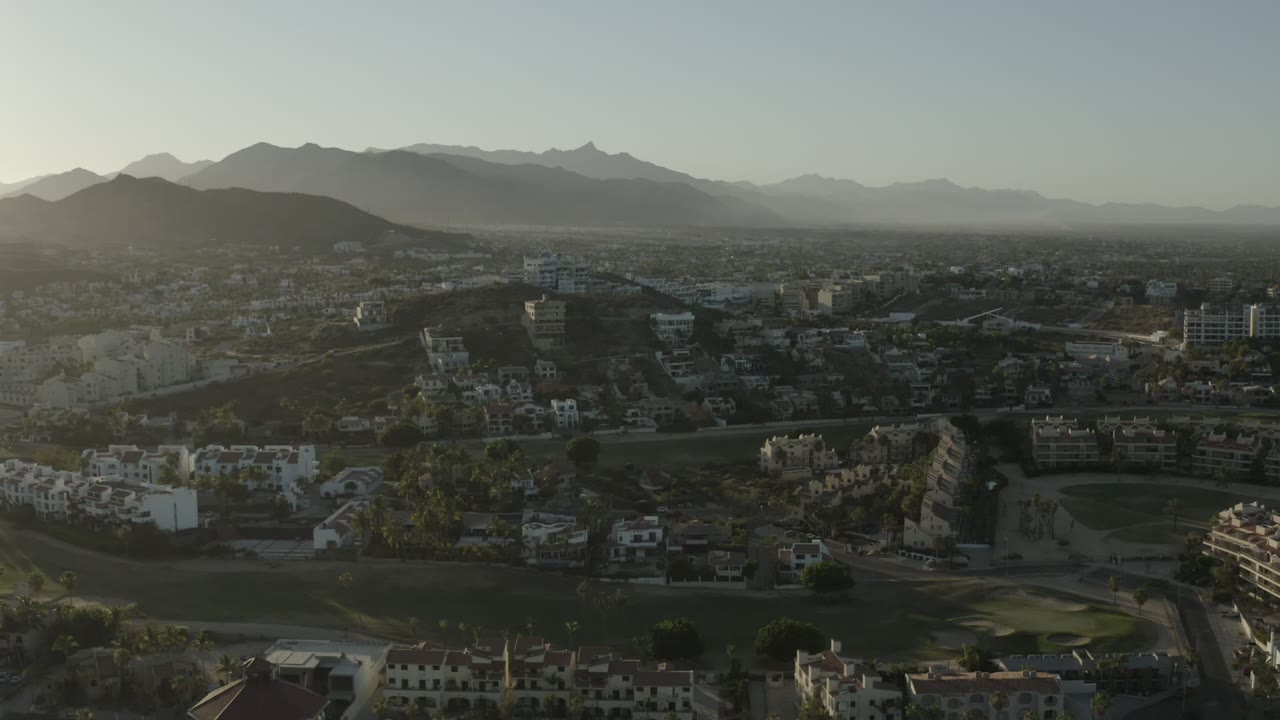 Second frame of the Los Cabos aerial view, with white residential blocks, a central road, landscaped green areas, and layered hills under hazy morning light, no visible people.