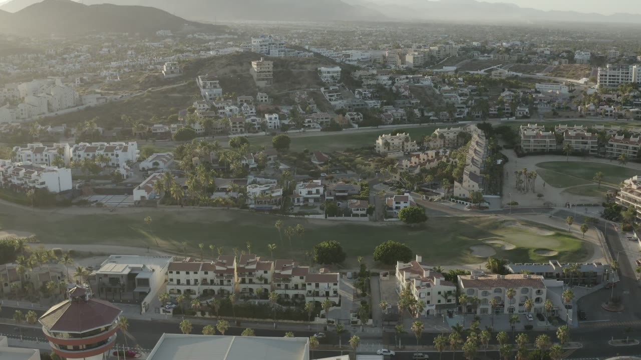 High aerial frame in Los Cabos showing dense residential blocks, a curving road, palm trees, golf-course landscaping, distant hills, and no clearly visible people.