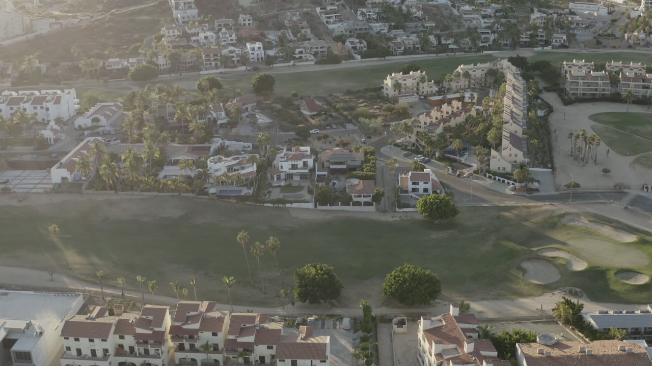 Aerial frame over Los Cabos, Mexico, showing low-rise homes, palm trees, a road, and a green grass field with sand bunkers, with no visible people.