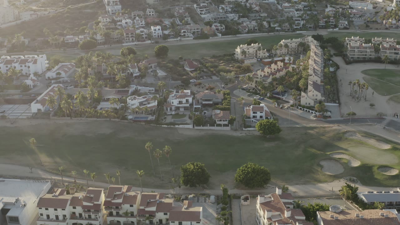 Drone view in Los Cabos, Mexico, with the field across the lower frame, houses and palms through the center, and hazy sunlight over the neighborhood, with no visible people.