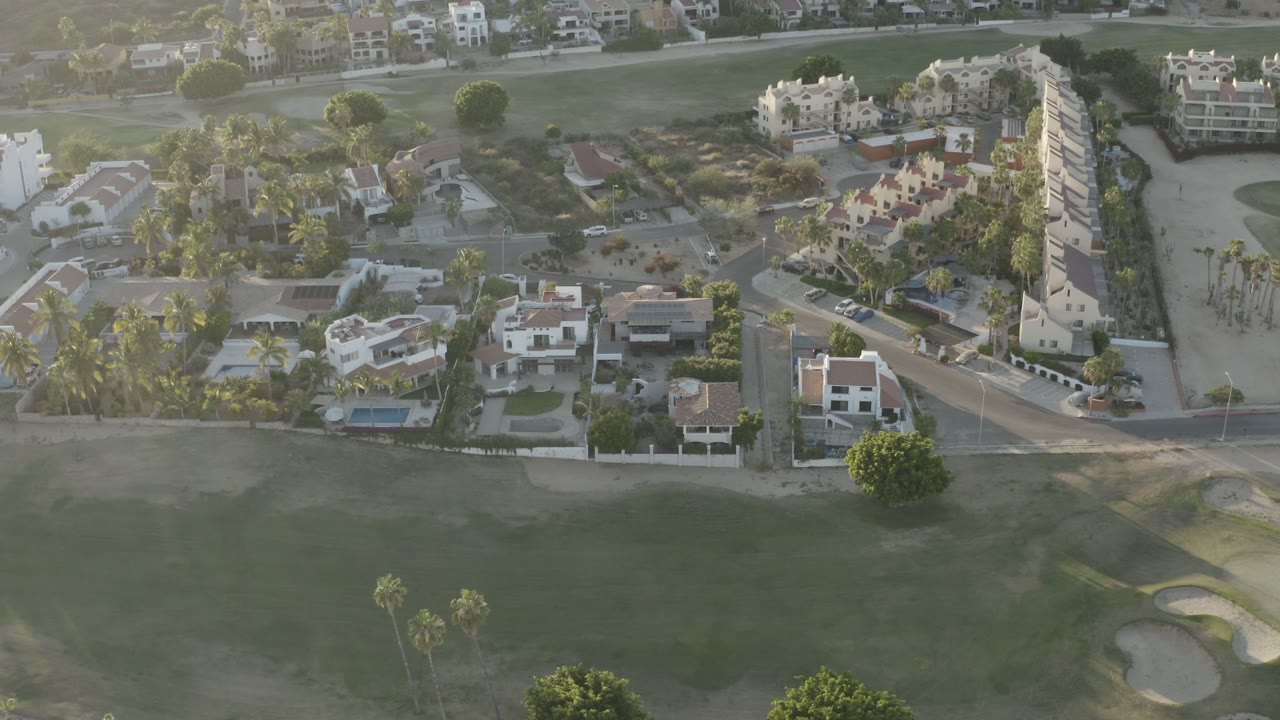 Aerial view in Los Cabos, Mexico showing houses, private pools, palms, a road, and golf course grass in warm light, with no visible people.