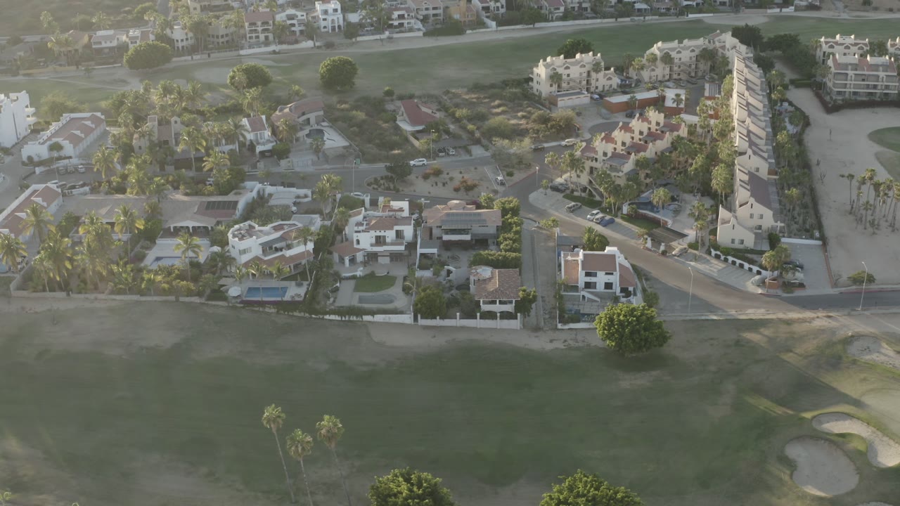 Aerial Los Cabos residential view with white homes, blue pools, palm-lined lots, a road crossing the frame, and golf course turf in the foreground, with no visible people.