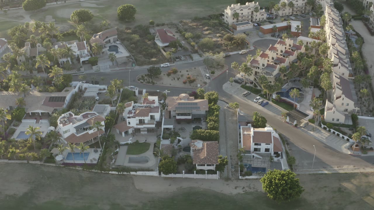 Aerial frame over Los Cabos showing clustered houses, green lawns, roadside parked cars, palms, and no visible people.