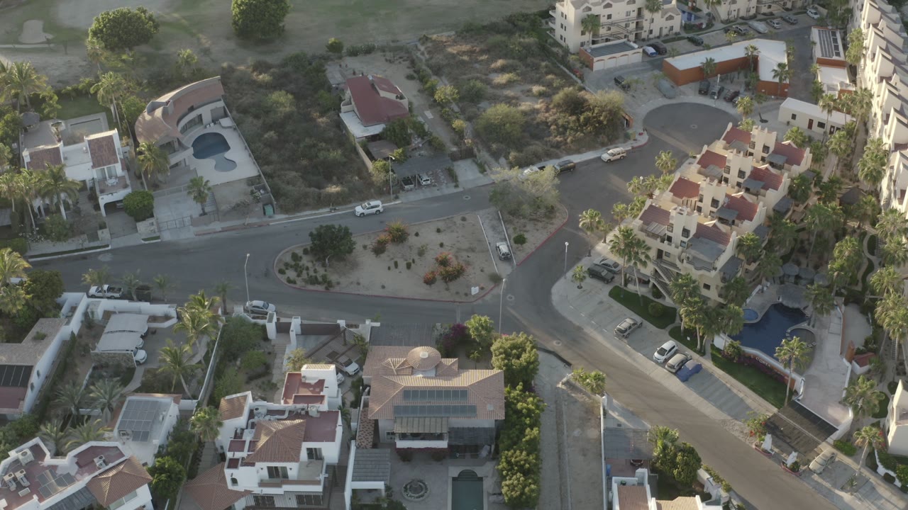 Overhead Los Cabos frame looking down on red-roofed homes, a curved road intersection, blue pools, palms, parked cars, and dry hillside brush with no visible people.