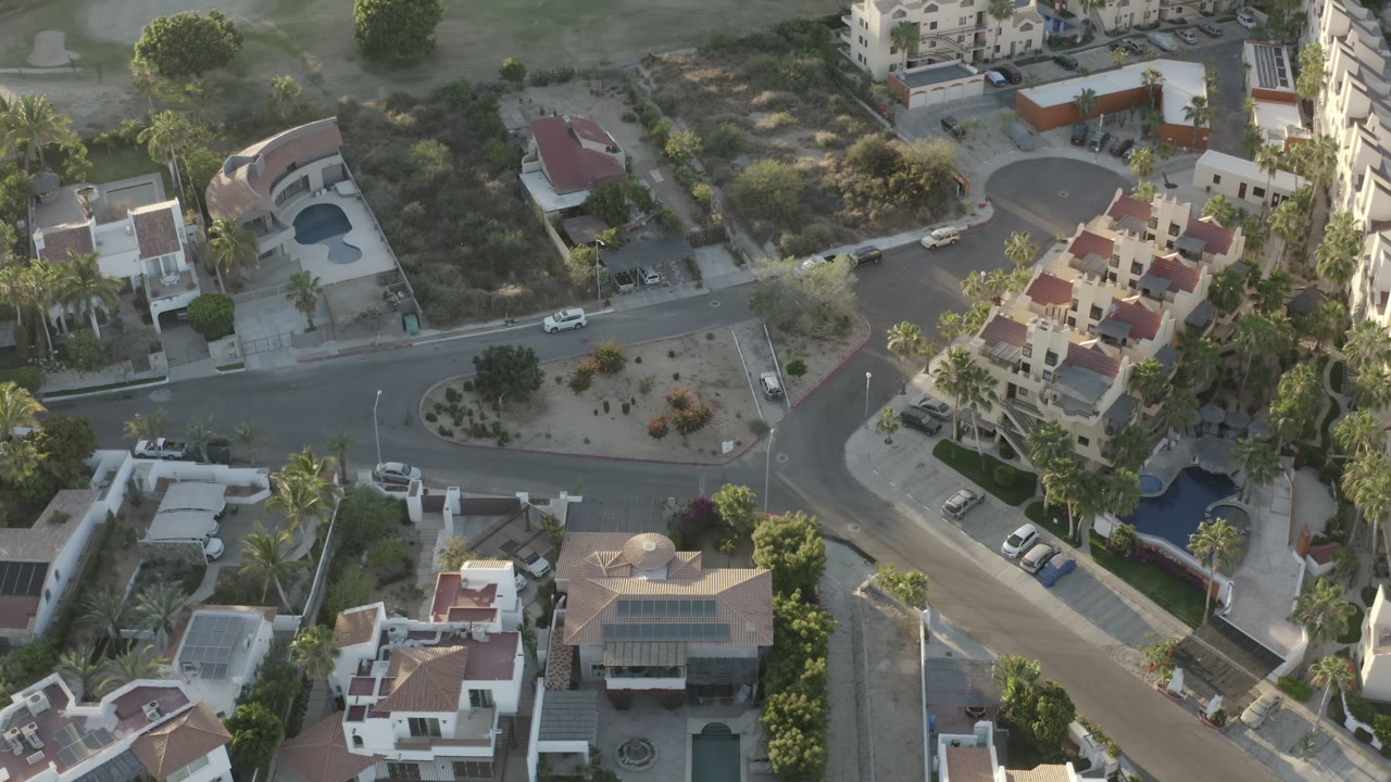 Bird's-eye Los Cabos view centered on a paved intersection, white and tan homes, red tile roofs, small blue pools, palms, and parked vehicles, with no visible people.