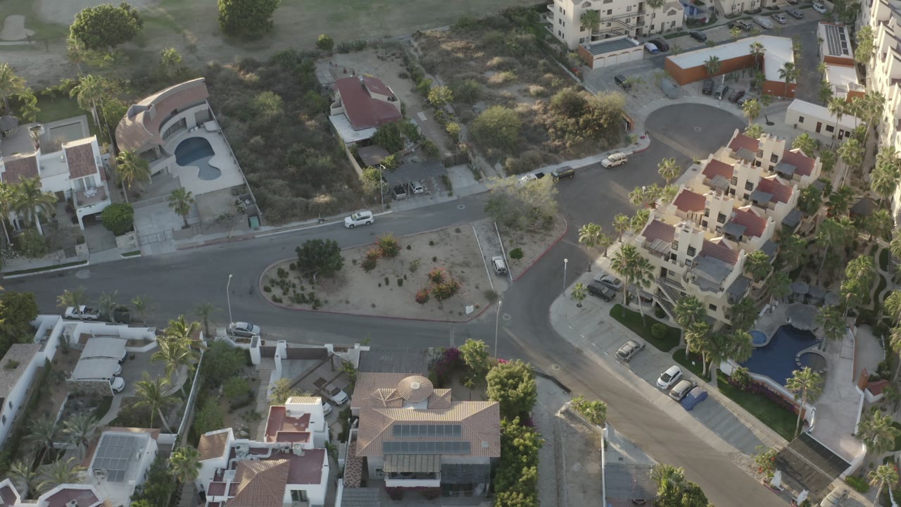 Aerial Los Cabos frame with a diagonal residential road, a small landscaped traffic island, private homes, pools, palm trees, and scrub-covered lots, with no visible people.