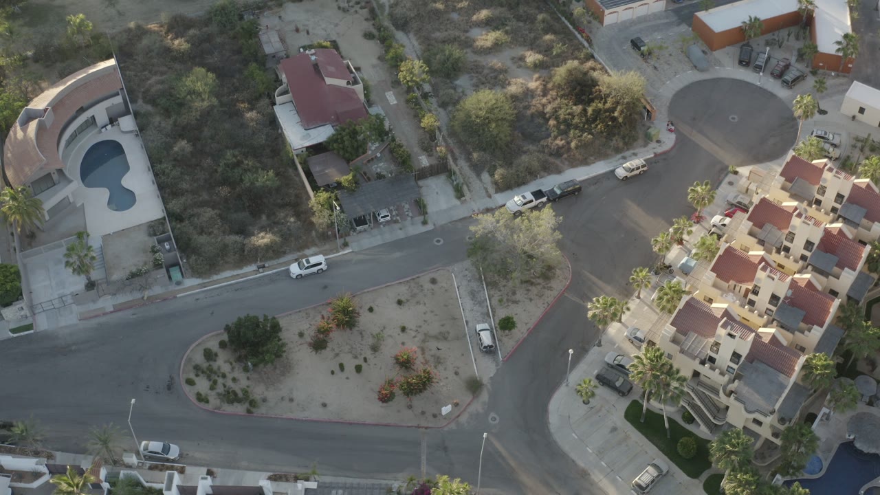 Overhead Los Cabos residential view with a curved pool at left, red-roofed homes at lower right, paved streets, parked cars, desert landscaping, and no visible people.