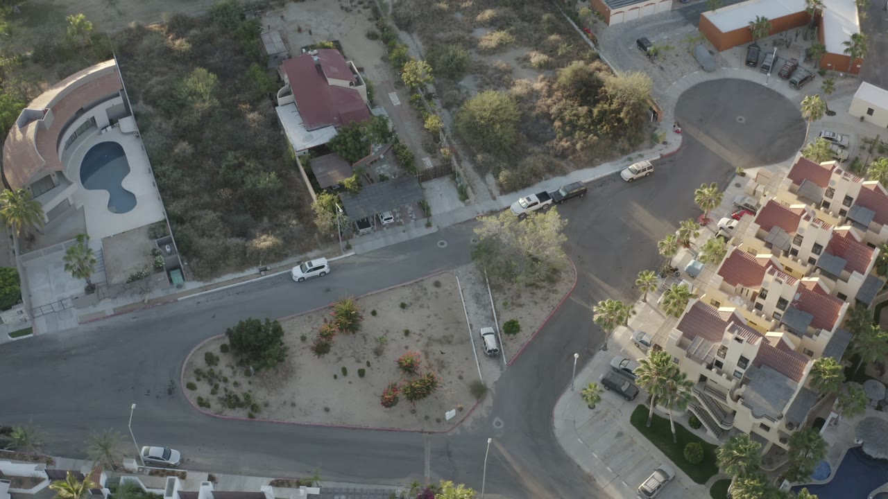 High overhead view in Los Cabos showing a road crossing, a circular cul-de-sac, red tile roofs, open desert lots, parked vehicles, and no visible people.