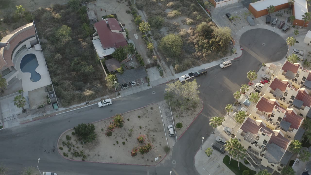 Top-down residential block with red-roofed homes, roads, vegetation, and parked cars.