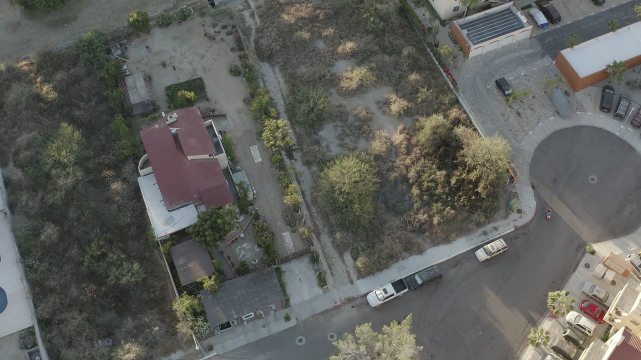 Top-down Los Cabos aerial frame showing a circular road end, dry brush, residential walls, small houses, parked vehicles, and no visible people.