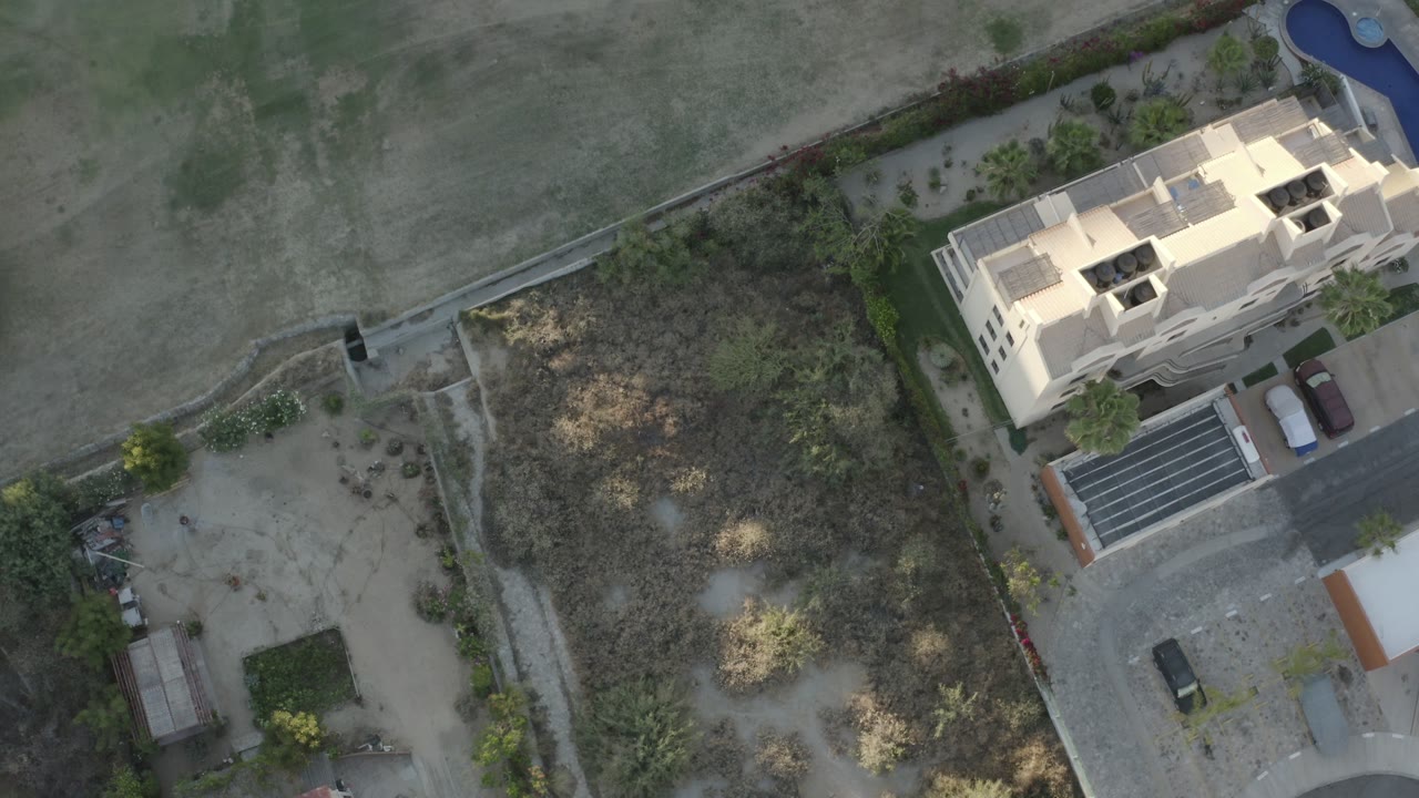 Aerial frame in Los Cabos, Mexico with the building still near the upper right, a dry field filling the upper half, scrubby vegetation in the middle, a driveway with vehicles, and no visible people.