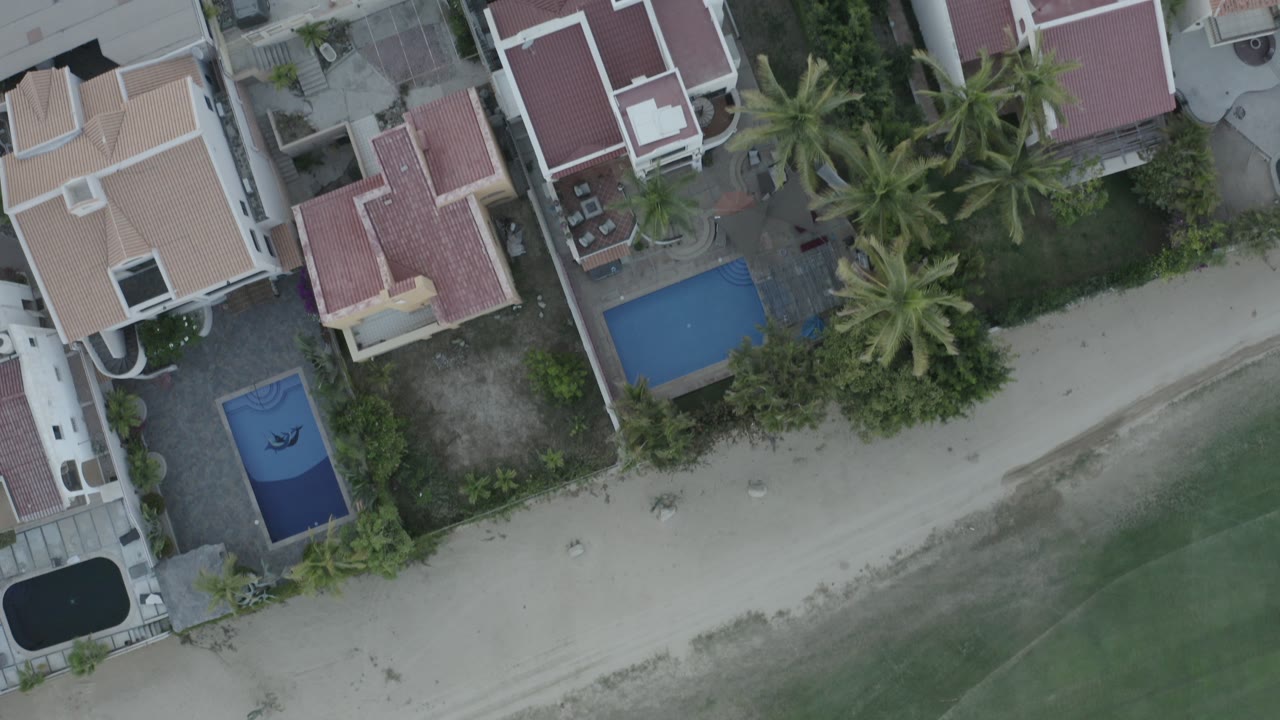 Top-down aerial frame over Los Cabos houses beside a sandy beach edge, with blue pools, palm trees, red and tan roofs, and no visible people.