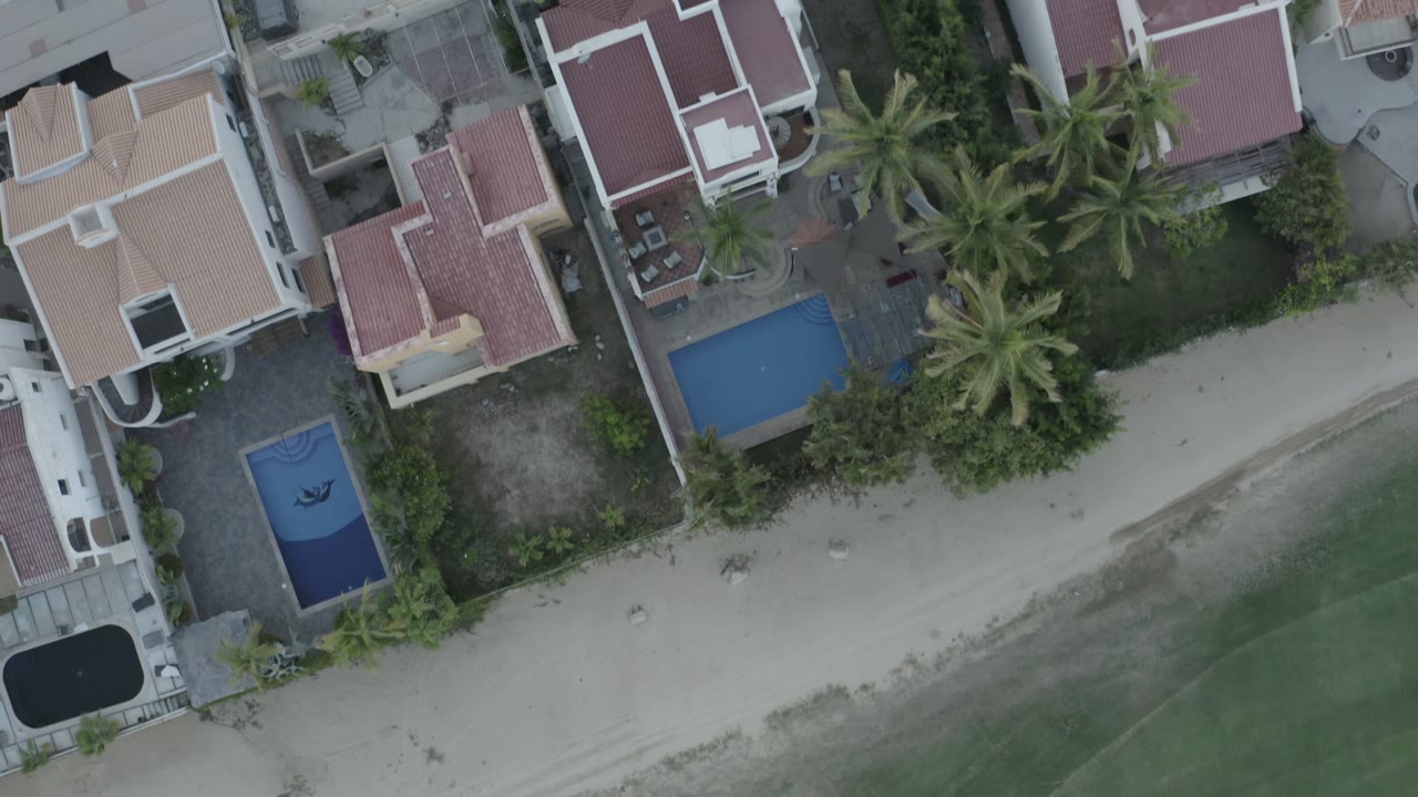 Los Cabos overhead view with rooflines, walled yards, palm clusters, a sandy shoreline, shallow green water, and no visible people.
