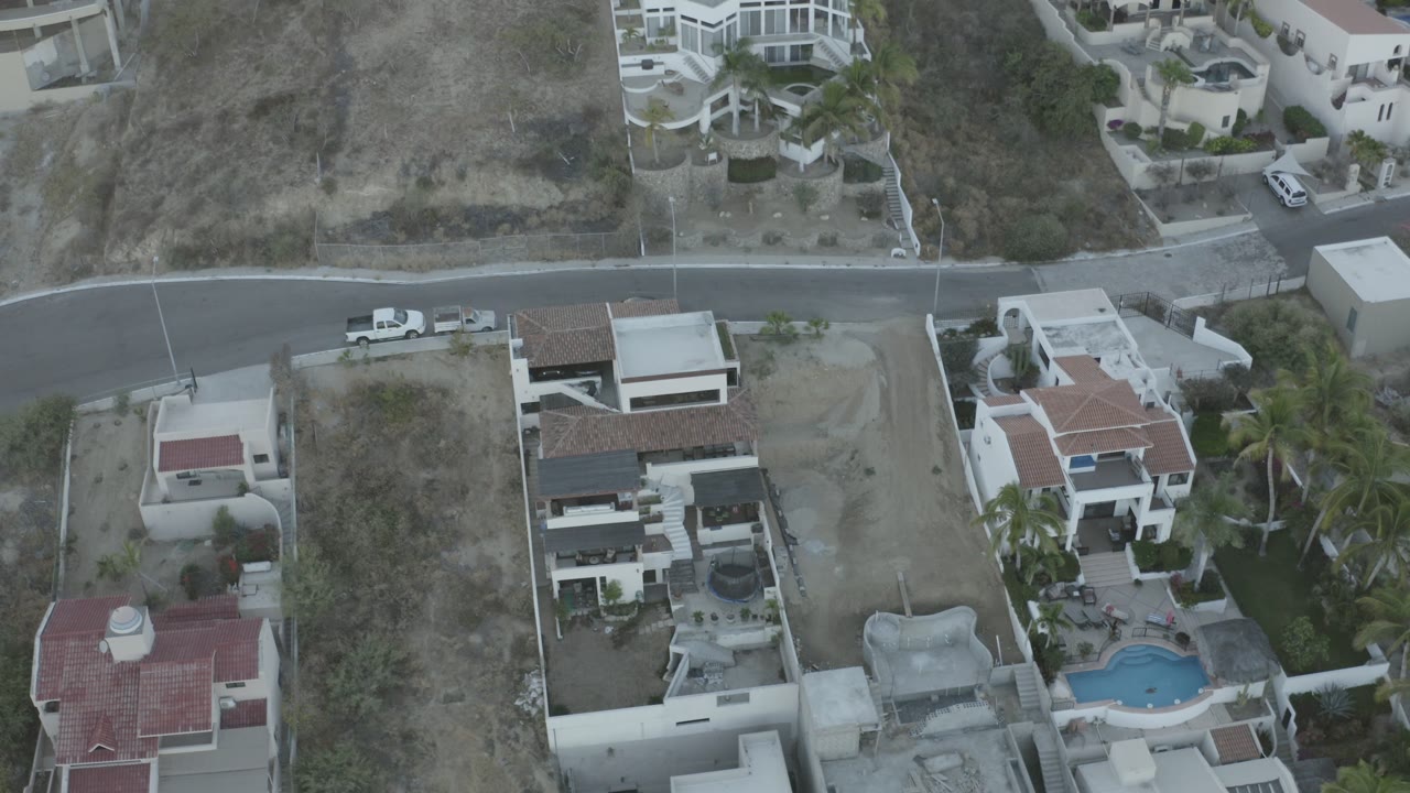 Aerial view over Los Cabos hillside homes, a curved road, parked vehicles, dry lots, and no visible people at the start of the clip.