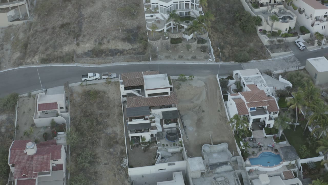 Drone view in Los Cabos looking down at white residential buildings, a dark curved street, hillside lots, parked vehicles, and no visible people.