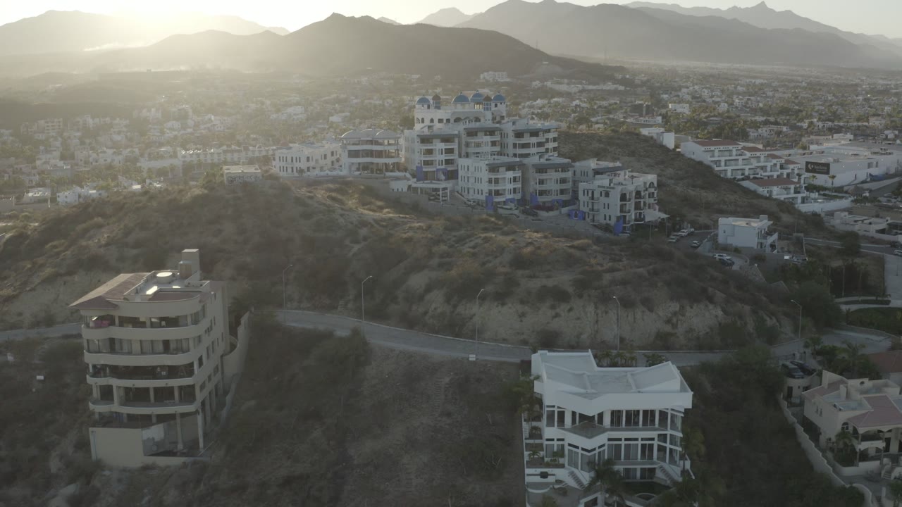 Los Cabos hillside view at the start of the clip, showing pale buildings, a road, dry slopes, hazy mountain ridges, bright backlight, and no visible people.