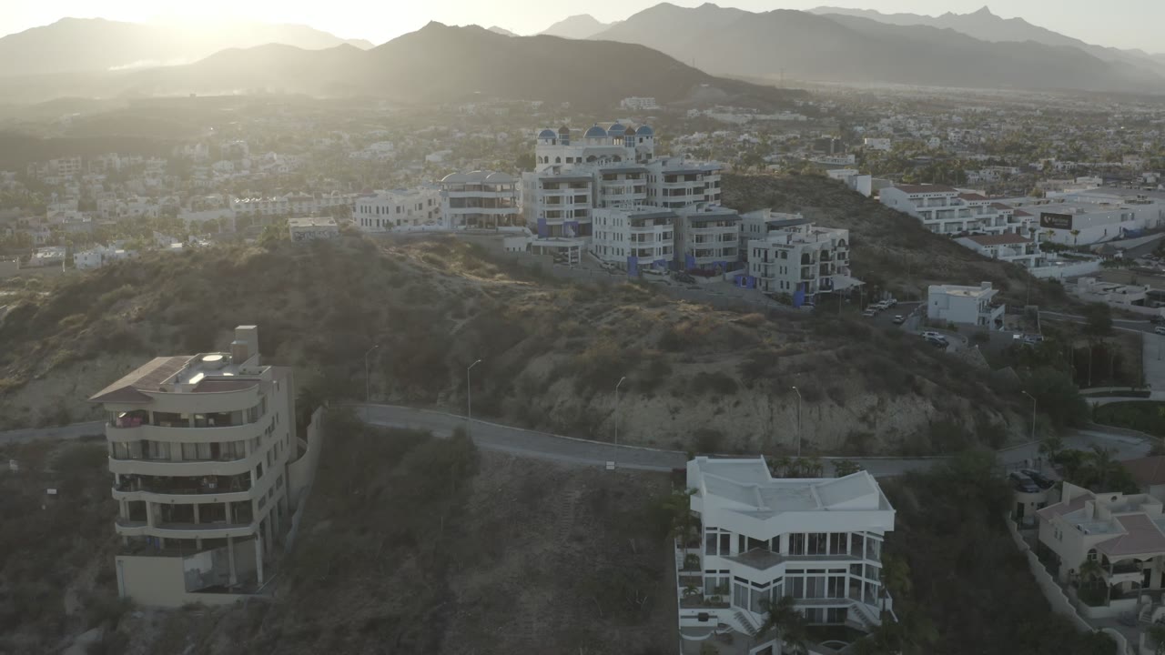 A slightly later Los Cabos frame shows the same hillside neighborhood, with a rounded building in the lower foreground, roads crossing the slope, hazy mountains, and no visible people.