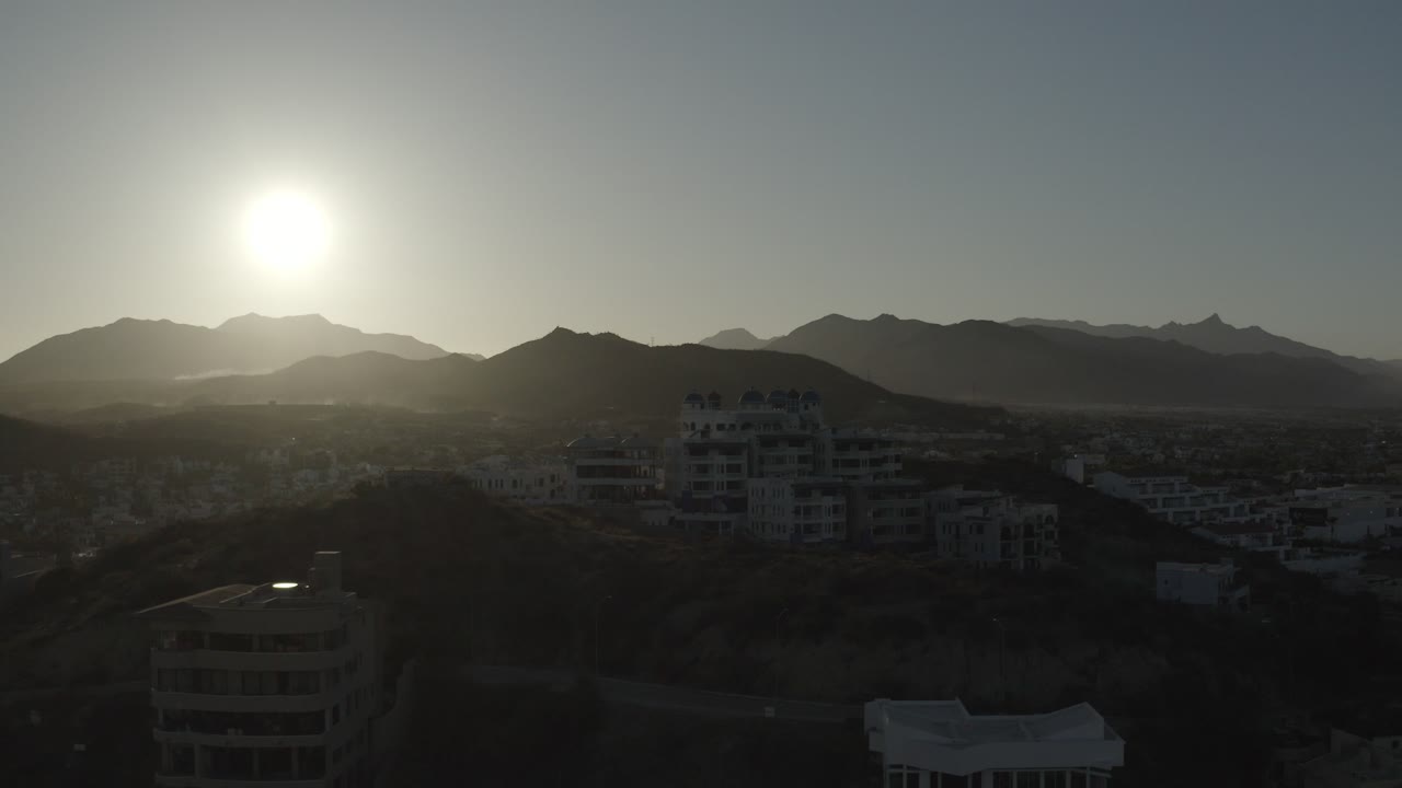 Los Cabos, Mexico sunrise frame with the sun above layered mountain ridges, dark foreground buildings, hazy morning light, and no visible people.