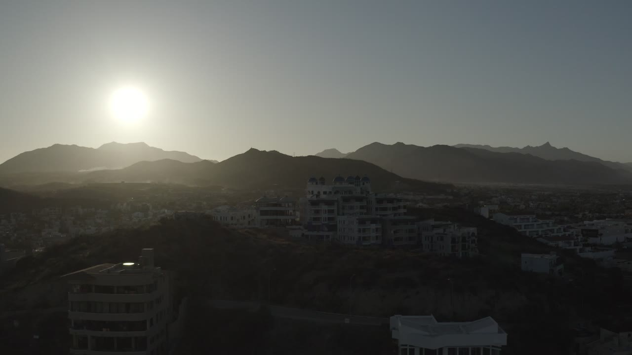 Los Cabos, Mexico view at 0:01 with the sun slightly left of center over mountain silhouettes, a dark hillside, clustered buildings, and no visible people.