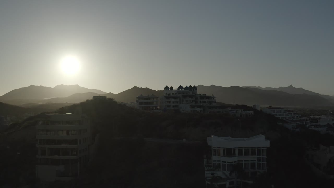 A low sun glows above dark residential buildings on a Los Cabos hillside, with layered mountain ridges behind and no visible people.