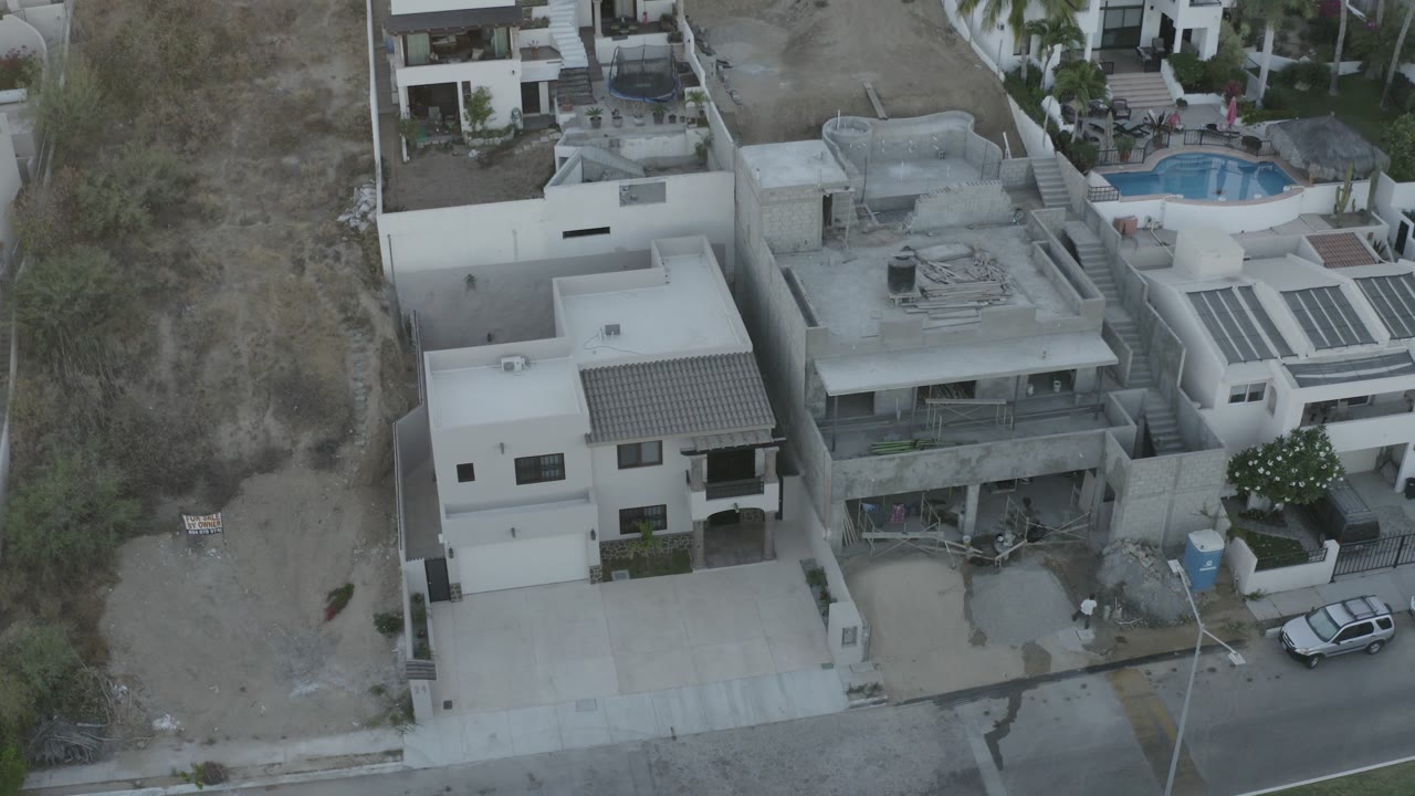 Second aerial frame in Los Cabos, Mexico holds on white residential buildings beside dry scrubland, with rooftop spaces, a blue pool beyond the homes, and a parked car along the street.