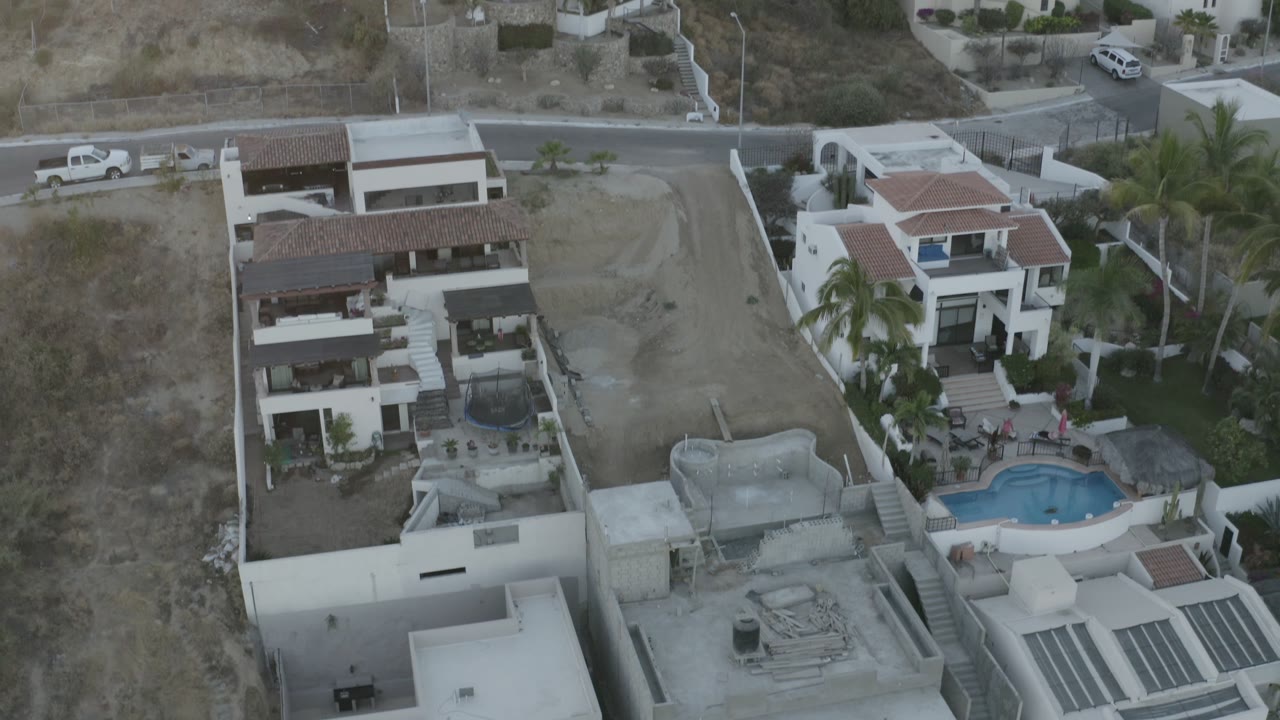 Side-top drone-style view in Los Cabos, Mexico, centered on hillside villas beside a curved road, with palms, walled lots, dry terrain, and no visible people.