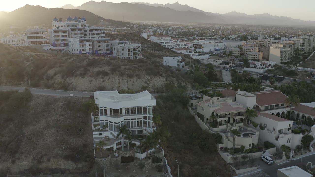 Backlit Los Cabos hillside with a large white house low in frame, tan homes beside it, ridge-top buildings, a spread of city blocks, mountains, and no visible people.