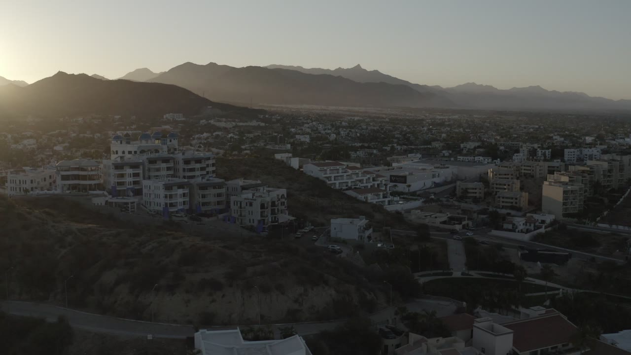 Aerial Los Cabos frame with low-rise buildings in shadow, a road crossing the lower hillside, hazy mountain ridges in back, and no visible people.