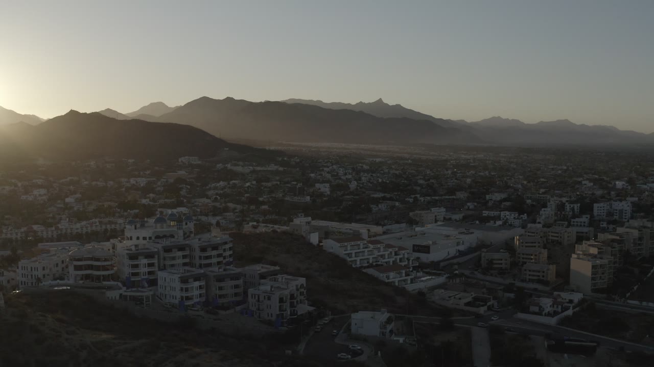 Aerial Los Cabos sunrise view with foreground apartment-style buildings, a dark hillside, a broad city grid, layered mountains, and no visible people.