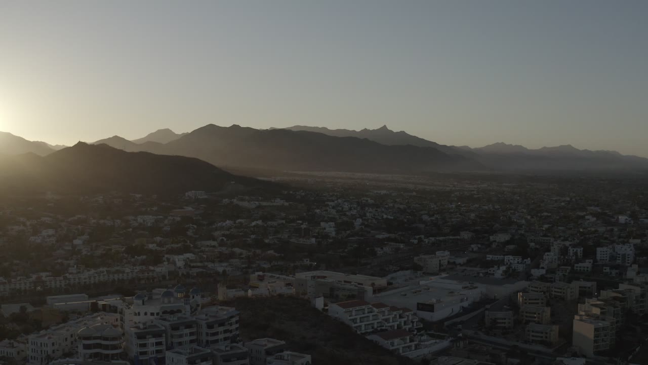 Los Cabos sunrise view with bright glare at the left edge, layered dark hills in the background, a dim urban foreground, and no visible people.