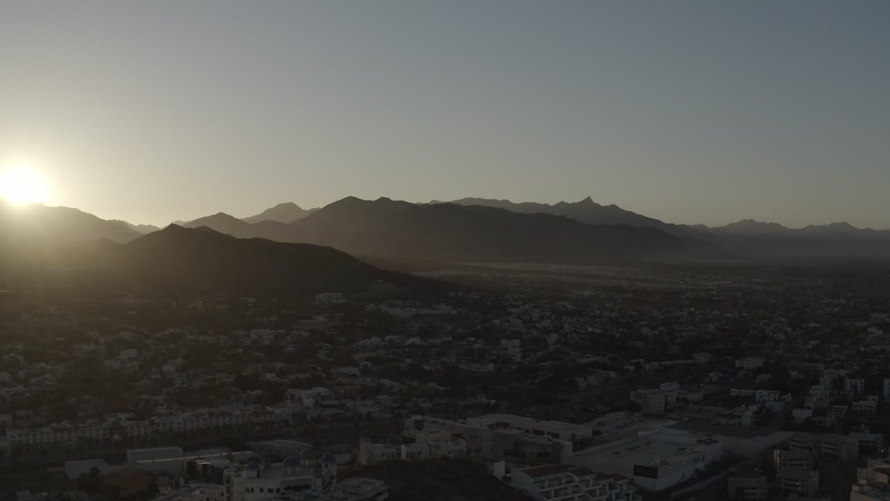 Sunrise view over Los Cabos with the bright sun still near the left edge, a broad mountain silhouette across the center, shadowed urban blocks below, and no visible people.