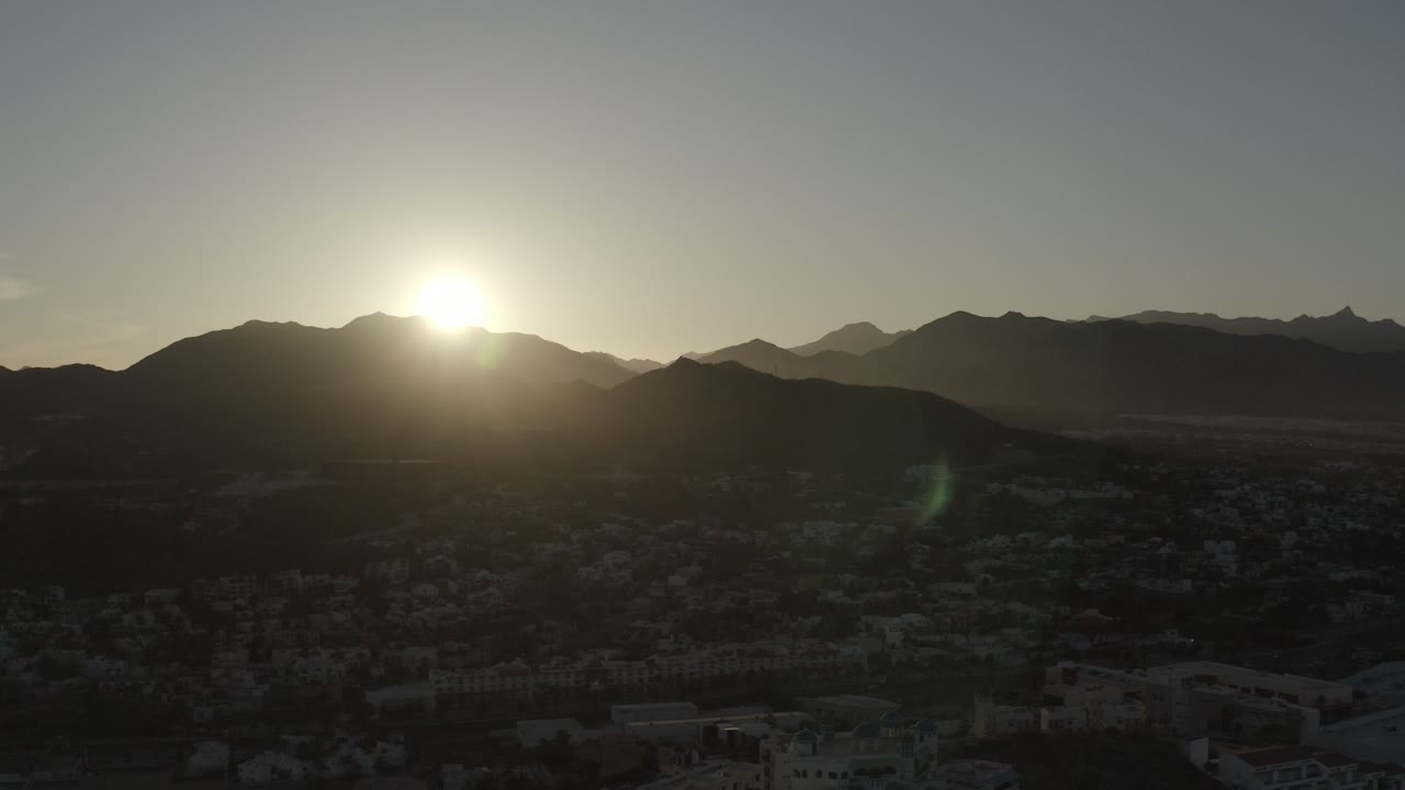 Low sun flares over a silhouetted mountain ridge near Los Cabos, Mexico, with houses in shadow below and no visible people.