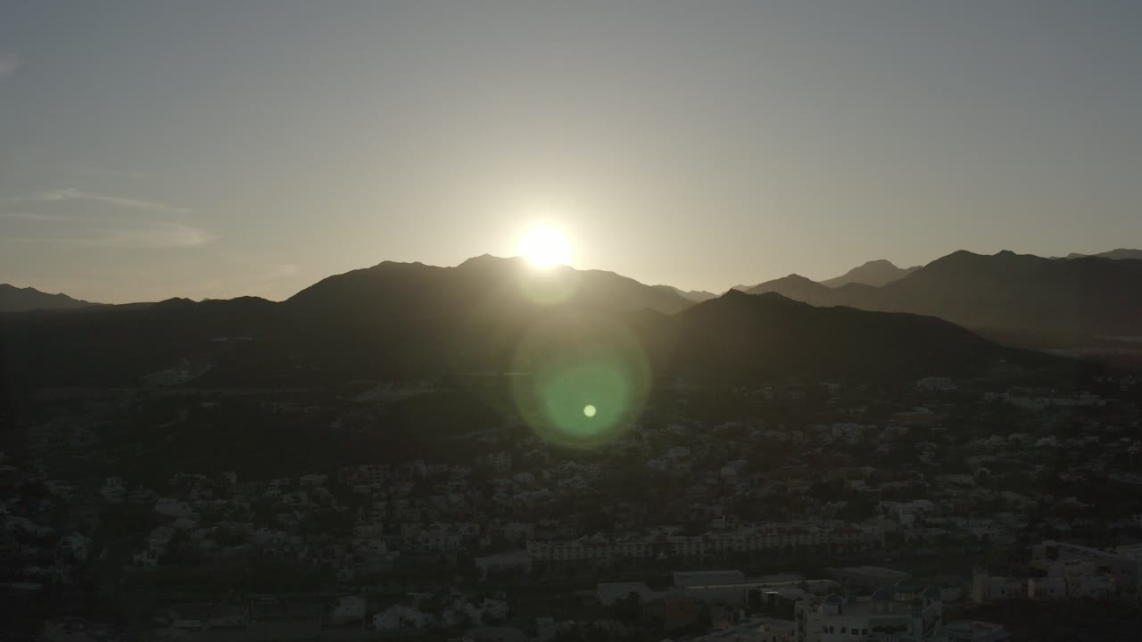 At 0:00, the Los Cabos sun sits just above a dark mountain ridge, with green lens flare over a shadowed town and no visible people.