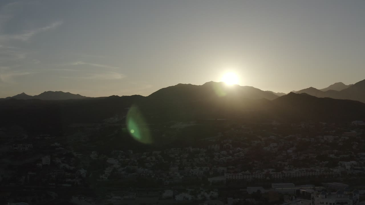 Los Cabos sunrise frame with the sun centered above the ridge line, layered mountain silhouettes, faint buildings in the valley, and no visible people.
