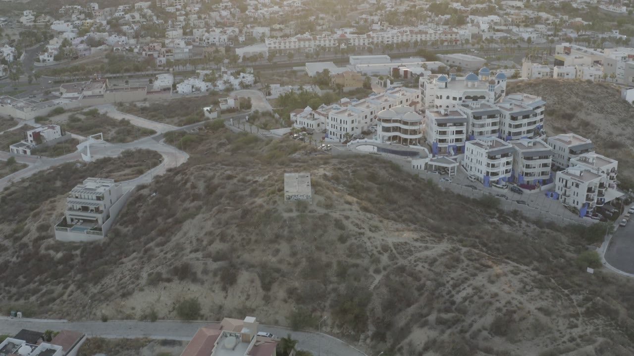 Elevated Los Cabos frame with a winding road around a brown hillside, white residential buildings in the midground, town blocks beyond, and no visible people.