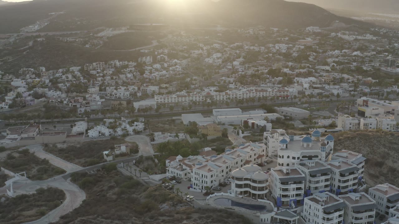 Second aerial frame over Los Cabos, Mexico, showing clustered light-colored buildings, a curved road at left, hazy hills, bright sun flare, and no visible people.