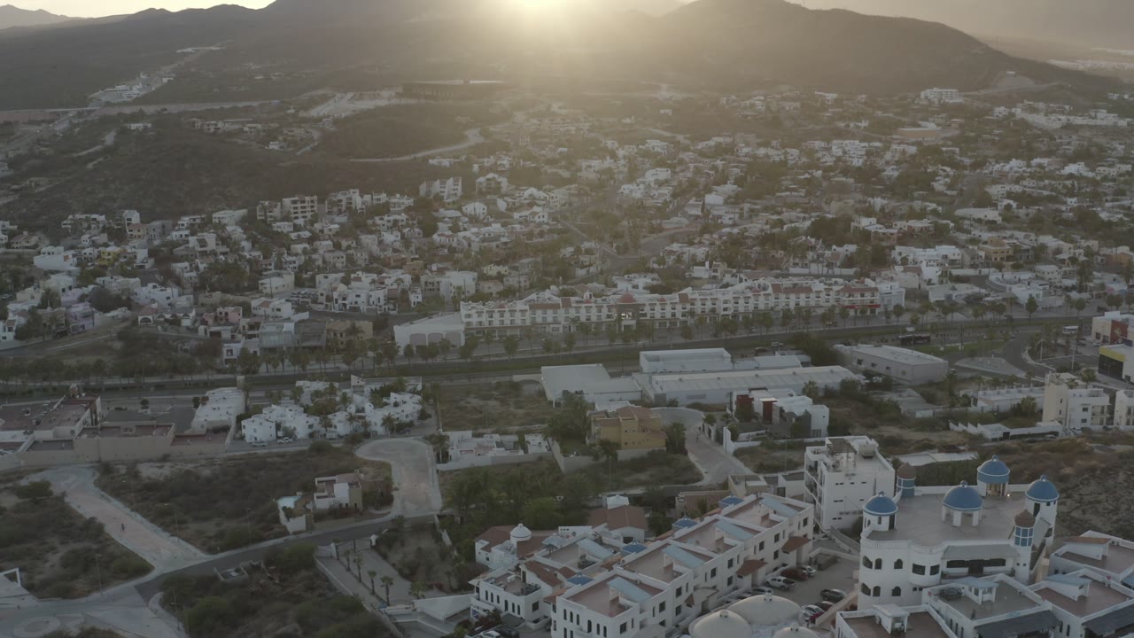 Aerial frame of Los Cabos hillside housing and roads, with a bright hazy sun above the mountain ridge and no visible people.