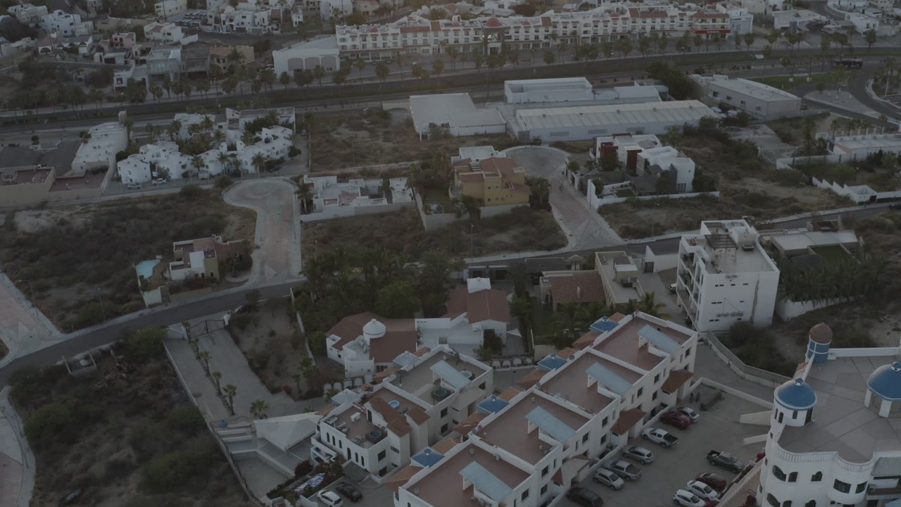 High aerial view over a Los Cabos residential block with white buildings, red tile roofs, parked cars, dry lots, and no visible people.
