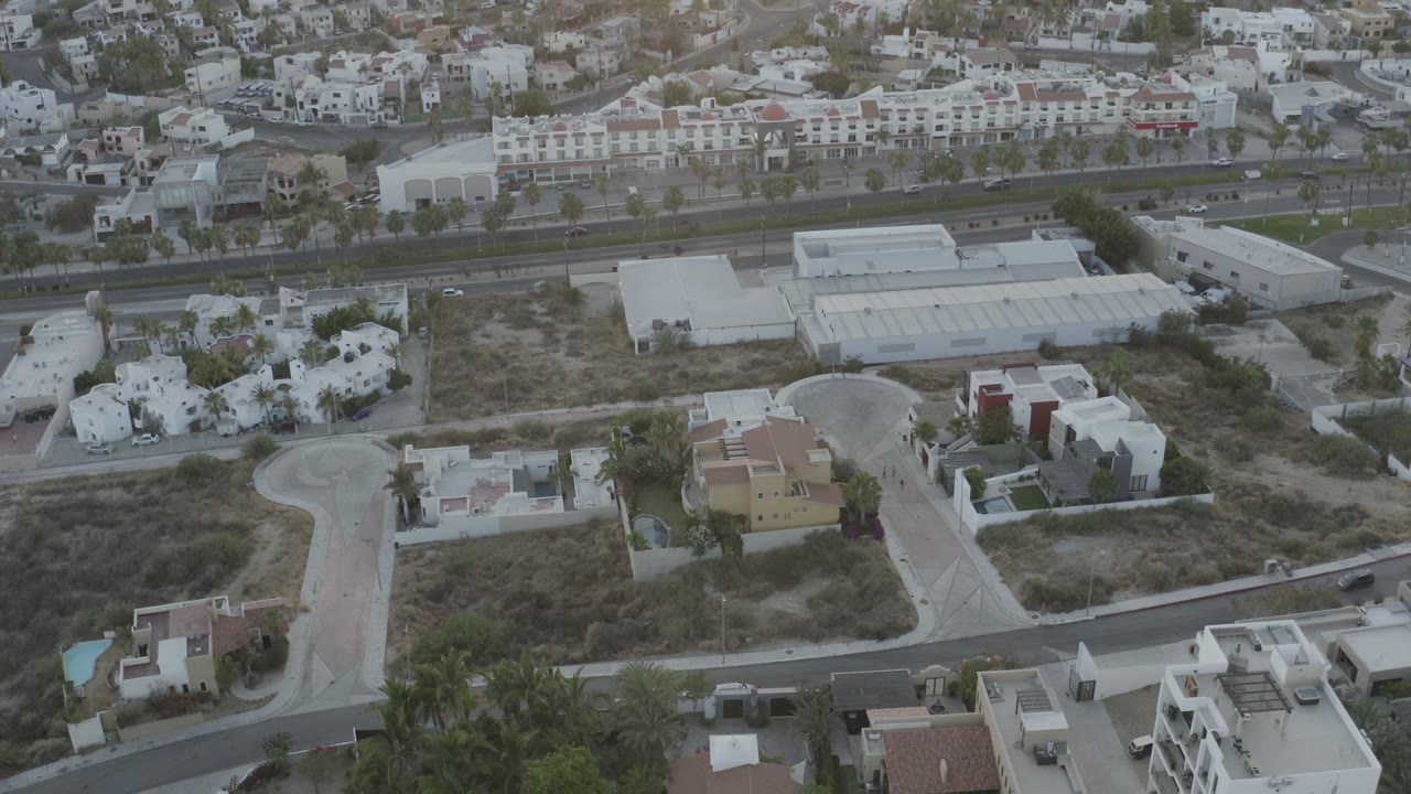 Opening aerial frame over Los Cabos, Mexico, showing pale industrial buildings, nearby homes, roads, scrub lots, palms, and no visible people.