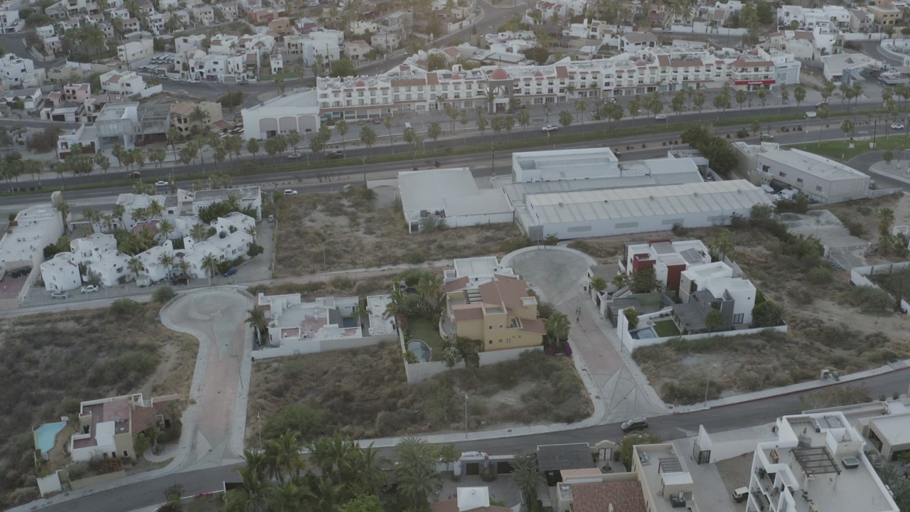 Aerial view in Los Cabos, Mexico, looking over sandy lots, white villas, a curving side street, and a wider road beyond, with no visible people.