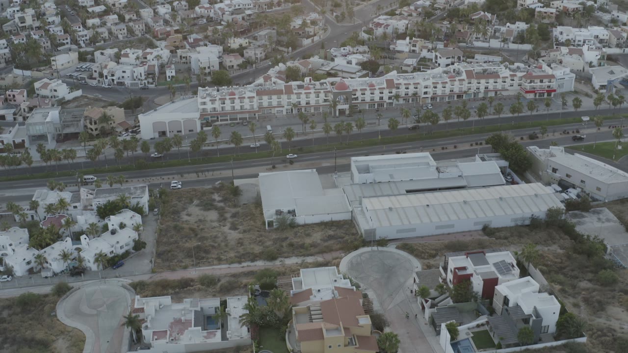 A slightly later aerial view in Los Cabos, Mexico, keeps the divided road centered with cars on the lanes, a large white-roofed building below, and dense residential buildings on the hillside, with no visible people.