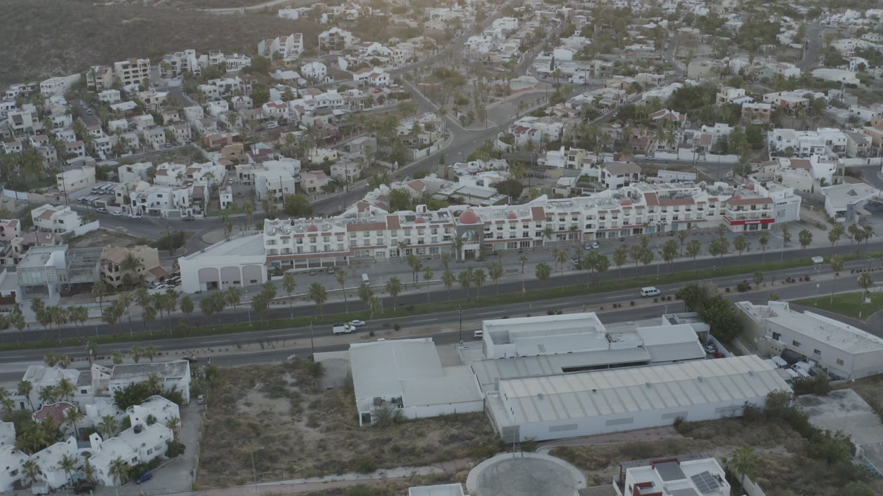 Opening aerial frame over Los Cabos, Mexico, showing white commercial buildings, a divided highway with small vehicles, hillside residences, and no visible people.